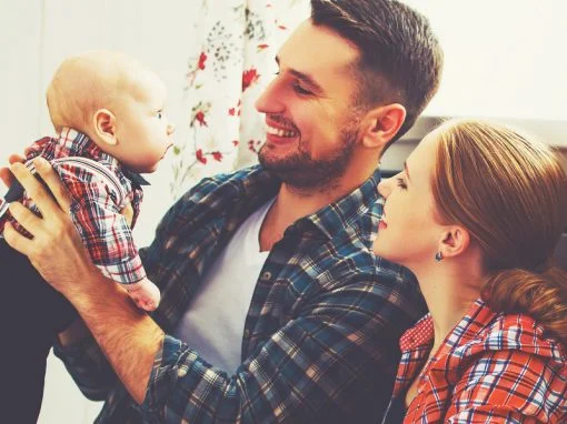 Young couple smiling as Dad holds young baby in the air, enjoying HCi cover » HCi