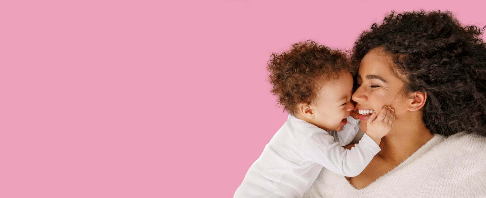 Smiling curly-haired woman with a baby grabbing her cheeks, on a pink background