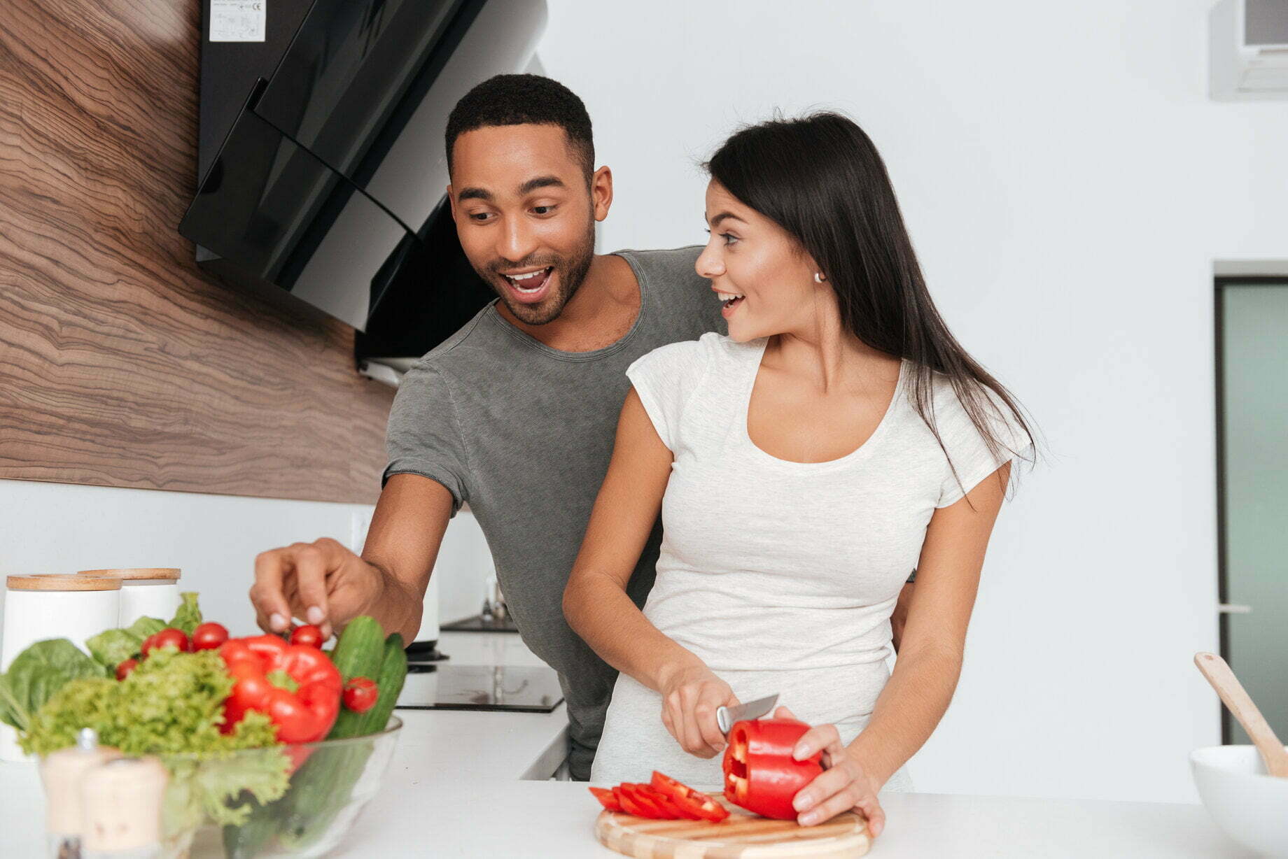 Smiling man takes a veggies from bowl beside woman cutting red capsicum