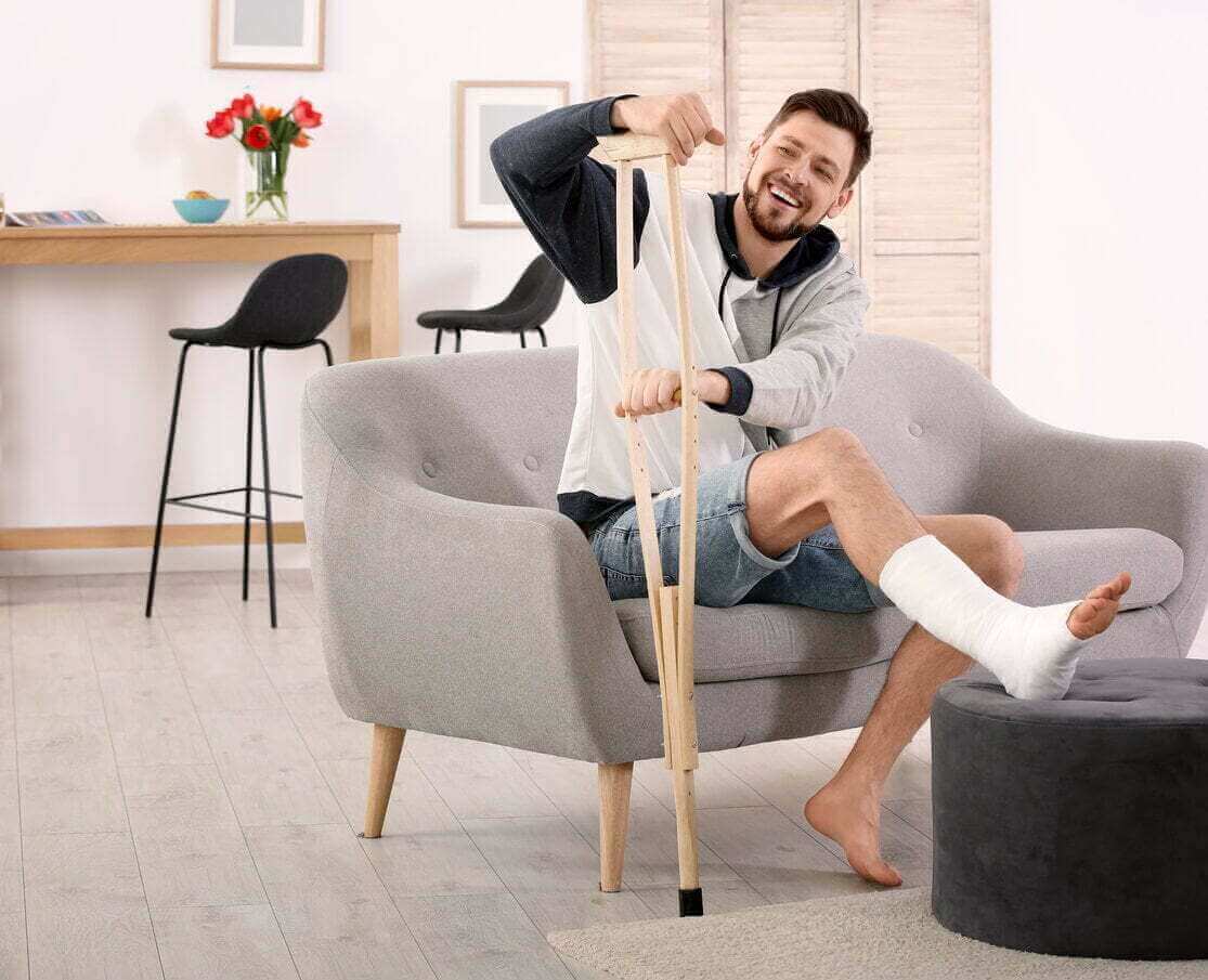 Smiling man with a bandaged ankle leaning on a crutch to get off the couch