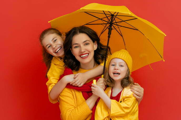 A smiling women with two young girls, all dressed in yellow rian coats and under a yellow umbrella