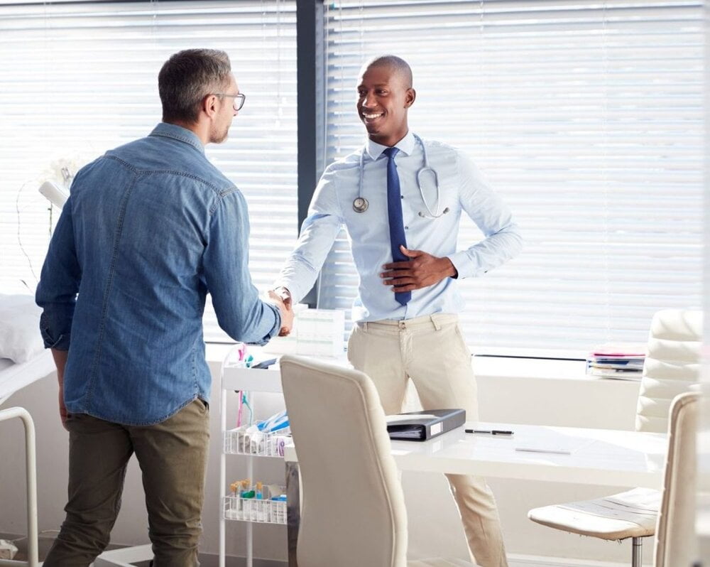 Doctor with stethoscope shaking hands with a patient over a desk before discussing a pre-existing condition and access gap costs » HCi